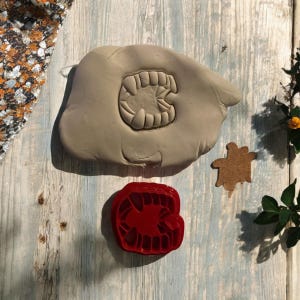 May include: A red plastic cookie cutter shaped like a set of fangs. The cookie cutter is on a wooden surface next to a piece of gray clay with an impression of the cookie cutter.
