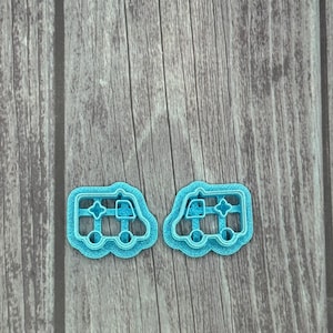 May include: Two light blue plastic cookie cutters shaped like ambulances. The cutters are on a woodgrain background.