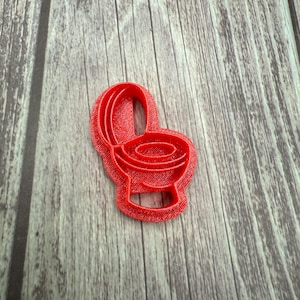 May include: A red plastic cookie cutter in the shape of a toilet. The cutter has a detailed design, showing the seat, bowl, and tank. The cutter is placed on a wooden surface.