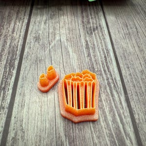May include: Orange plastic cookie cutters shaped like a popcorn bucket and two popcorn kernels.