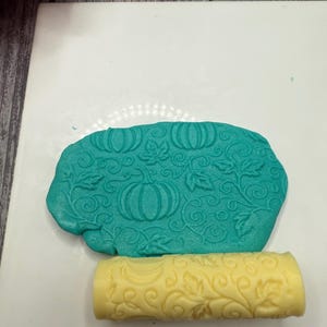 May include: A teal clay piece with embossed pumpkins and leaves, alongside a yellow roller with a similar leaf pattern. The items are on a white surface, suggesting a crafting or baking theme. The roller is used to create the embossed design.