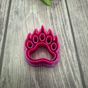 May include: A pink plastic bear paw cookie cutter. The cutter has five rounded holes for the toes and a raised edge around the paw print.