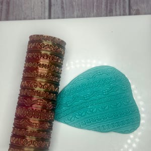 May include: A stack of decorative, ornate bangles in shades of brown and green, with intricate floral and geometric patterns. A turquoise, leaf-shaped item with a detailed design is next to the bangles.