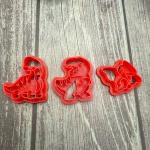 May include: Three red plastic dinosaur-shaped cookie cutters. The cutters are in different poses, including a standing dinosaur, a dinosaur with its mouth open, and a dinosaur head. The cutters are on a wooden surface.