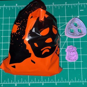 May include: A black and orange plastic bag with a black and white graphic design. The bag is on a green cutting mat with two purple plastic cookie cutters. One cookie cutter is a three-leaf clover shape and the other is a round shape with a design.