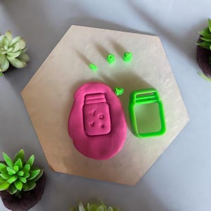 May include: A green plastic cookie cutter shaped like a mason jar with a lid. The cookie cutter is on a white surface with a pink piece of play dough that has been pressed with the cookie cutter.