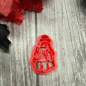 May include: A red plastic cookie cutter in the shape of a person wearing a hat and holding a mug. The cutter is on a wooden surface with red glittery flower petals.