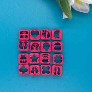 May include: Sixteen pink cookie cutters in various shapes, including a flower, heart, rainbow, star, and four-leaf clover. The cutters are arranged in a grid on a blue surface, with a white flower and green stem in the background.