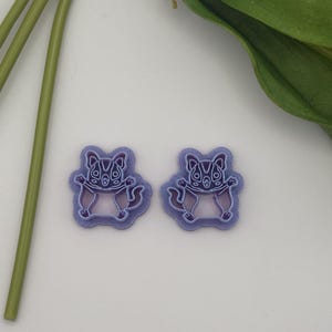 May include: Two purple and lavender bat-shaped craft embellishments. Each bat has a detailed design with a light purple body and darker purple accents. The bats are on a light background, with green stems and leaves in the background.