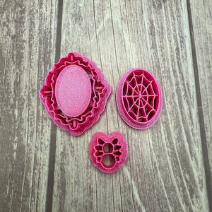May include: Three pink cookie cutters on a wooden surface. The cutters include an oval frame, a spiderweb, and a cat face. The cutters are made of plastic and are designed for baking. The background is a light brown wood.