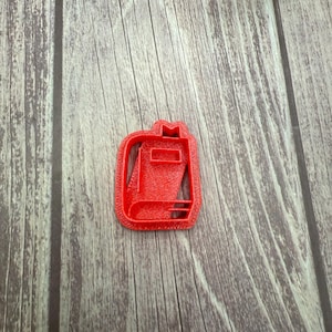 May include: A bright red cookie cutter in the shape of a backpack with a book inside. The cutter is made of plastic and sits on a wooden surface.