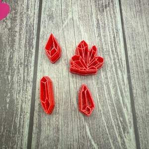 May include: Four red plastic cookie cutters shaped like crystals. The cutters are on a wooden surface.