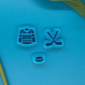 May include: Three blue plastic cookie cutters in the shape of hockey-themed items. The shapes include a hockey jersey, hockey sticks, and a hockey puck. The items are on a blue background.