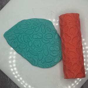 May include: A teal clay sheet and a red roller with a nut pattern. The clay sheet is imprinted with various nut shapes, including peanuts and walnuts. The roller has a similar design, perfect for creating textured surfaces.