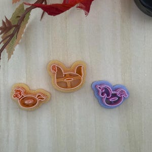 May include: Three plastic cookie cutters in the shapes of a chicken, a rooster, and a unicorn. The chicken and rooster cutters are orange, while the unicorn cutter is purple. Each cutter features a raised outline and a central detail design.