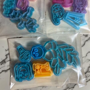 May include: A collection of plastic cookie cutters in various shapes including a dragon, a sewing machine, a skull, a leaf, and character faces, packaged in clear plastic bags. The cutters are in shades of blue, yellow, pink, and purple.