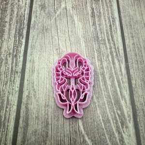 May include: A pink plastic cookie cutter in the shape of a stylized floral design.