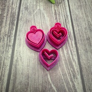 May include: Three pink heart-shaped cookie cutters of different sizes. The cutters are made of plastic and have a shiny finish.