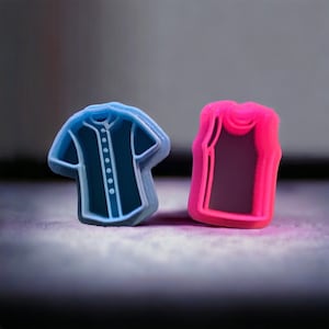 May include: Two small, 3D printed plastic figurines of clothing items. The figurine on the left is a light blue baseball jersey. The figurine on the right is a pink tank top.