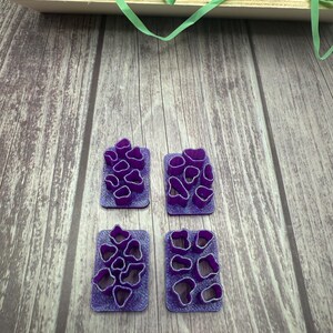 May include: Four purple plastic cookie cutters with different shapes of flowers. The cutters are on a wooden surface.