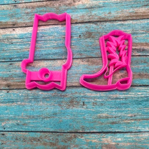 May include: Two pink plastic cookie cutters, one shaped like a cowboy boot and the other shaped like a hat.