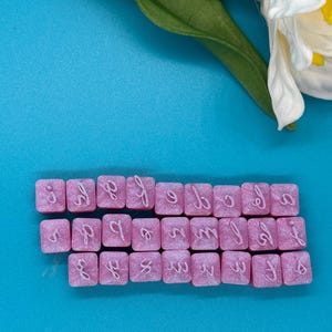 May include: Pink resin alphabet letter tiles with white cursive lettering, arranged on a blue surface. A green stick and a white and yellow flower are in the background. The tiles are square and have a glittery finish.