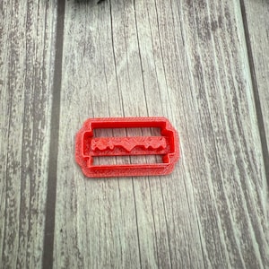 May include: A red plastic cookie cutter in the shape of a razor blade. The cutter has a rectangular frame with a detailed blade design inside. The cutter is on a wooden surface.