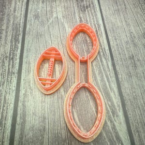 May include: Two pink plastic cookie cutters shaped like a football. The football cutter has a detailed design of laces and stitching.