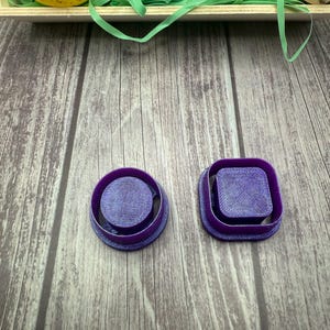 May include: Two purple plastic cookie cutters, one round and one square, on a wooden surface.
