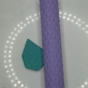 May include: A lavender-colored, cylindrical object with a scale pattern, next to a teal, teardrop-shaped object with a matching scale pattern. The objects are likely decorative or craft supplies.