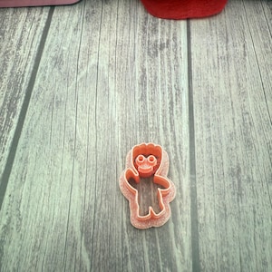May include: A pink plastic cookie cutter shaped like a cartoon character with a big head, two eyes, and a smile. The cookie cutter is on a wooden surface.