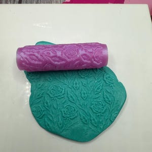 May include: A purple roller with a floral rose pattern rests on a teal clay piece, also with a rose and leaf design. The roller is cylindrical, and the clay is a rounded shape. The background is a white surface.