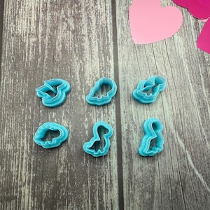 May include: Six light blue plastic dinosaur-shaped cookie cutters are arranged on a wooden surface. The cutters are in various poses, including a T-Rex and a Brontosaurus. Pink and red paper hearts are in the background.