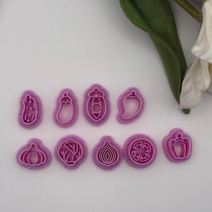 May include: A set of ten small, pink vegetable-shaped cookie cutters. The shapes include a carrot, eggplant, pepper, chili pepper, garlic, cabbage, onion, tomato, and bell pepper. The cutters are arranged on a white surface.
