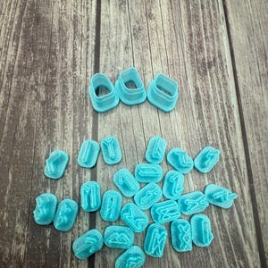 May include: A set of 28 blue plastic cookie cutters shaped like the runes of the Elder Futhark alphabet. The cutters are arranged in a scattered pattern on a wooden surface. There are also three larger blue plastic cookie cutters in the shape of a rectangle.