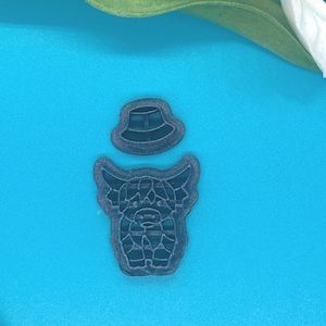 May include: Two dark gray cookie cutters: one shaped like a Highland cow and the other a small hat. The cow cutter has detailed features, including horns and a textured body. The hat cutter is a simple, rounded design. Both are on a blue surface.