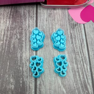 May include: Four light blue heart-shaped cookie cutters. The cutters are arranged on a wooden surface. A pink box and red heart-shaped cutouts are visible in the background. The cutters are for baking and crafting.