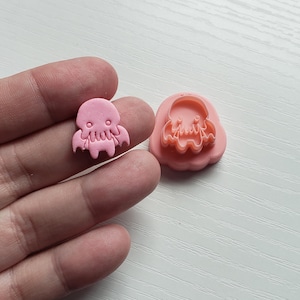 May include: Pink plastic cookie cutter in the shape of a cartoon Cthulhu with a toothy grin. The cutter is shown next to a pink Cthulhu cookie.