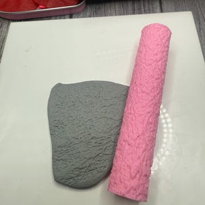 May include: A pink textured roller and a gray textured piece of clay on a white surface.