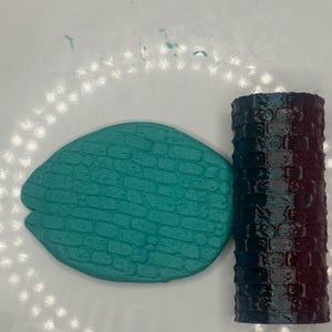 May include: A teal-colored, oval-shaped item with a brick pattern next to a cylindrical object with a brick pattern that fades from teal to burgundy. The objects are likely handmade and could be used for crafts or home decor.