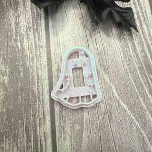 May include: A pink plastic cookie cutter in the shape of a ghost with a light blue outline. The ghost has a rectangular opening in the center and decorative cutouts. A black fabric flower is in the background.