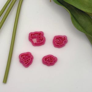 May include: Four pink cookie cutters in various animal shapes, including a pig, a fish, and a turtle. The cutters are on a white surface, with green stems and leaves in the background.