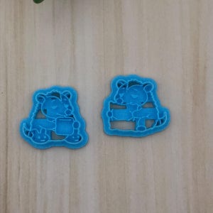 May include: Two bright blue plastic cookie cutters featuring a cartoon character design. The left cutter depicts a character holding a cup, while the right cutter shows a character leaning against a horizontal bar. Both have a textured finish.