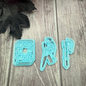 May include: Three light blue cookie cutters in the shapes of a book, a person, and an axe. The cutters are made of plastic and are set against a wooden background. The book cutter has a detailed design.