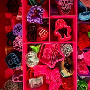 May include: A pink plastic storage case filled with various colorful plastic cookie cutters. The cutters are shaped like ghosts, pumpkins, bats, and other Halloween themes.