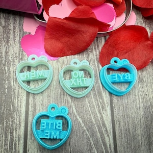 May include: Four heart-shaped cookie cutters in shades of blue and teal, each with a different word or phrase: "WEH", "XHT", "BAE", and "BITE ME". Red and pink heart-shaped confetti and rose petals surround the cutters on a wooden surface.