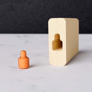 May include: An orange, textured bottle with a rounded top sits next to a rectangular, cream-colored block with a bottle-shaped cutout. The objects are on a white marble surface.