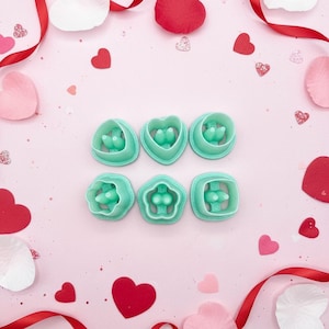 May include: Six teal-colored cookie cutters in various shapes, including hearts, circles, and a flower, are arranged on a pink surface. The cutters have a bird-like design inside. Red heart-shaped confetti and ribbon, along with white petals, surround the cutters.