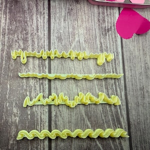 May include: Four yellow, wavy, decorative ribbons are arranged on a wooden surface. The ribbons have a soft, textured appearance and are in various shapes. A pink heart shape is visible in the background.