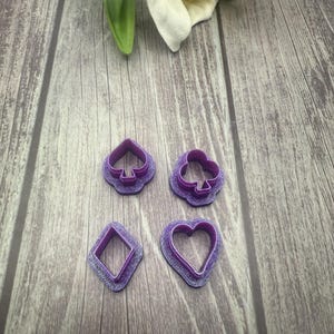 May include: Four purple plastic cookie cutters shaped like a heart, a diamond, a spade, and a club.